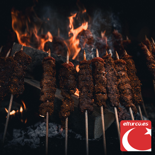 Brochetas de carne picada asándose sobre las brasas, con llamas al fondo