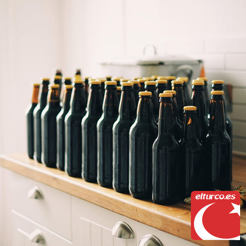 Filas de botellas de cerveza oscuras sin etiqueta sobre una encimera de madera, con chapas doradas al lado