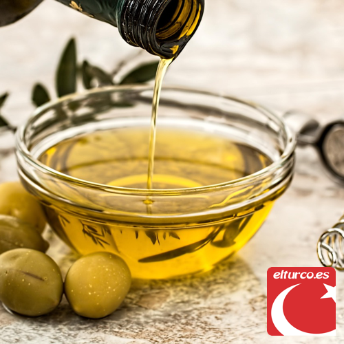 Aceite de oliva virgen vertido desde una botella en un cuenco de cristal, junto a aceitunas verdes